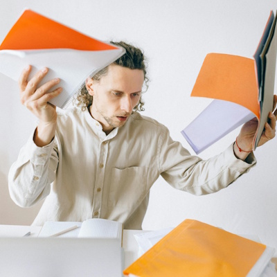 A person with curly hair wearing a beige shirt appears overwhelmed while holding several colorful folders in both hands, surrounded by more folders and papers on a table.