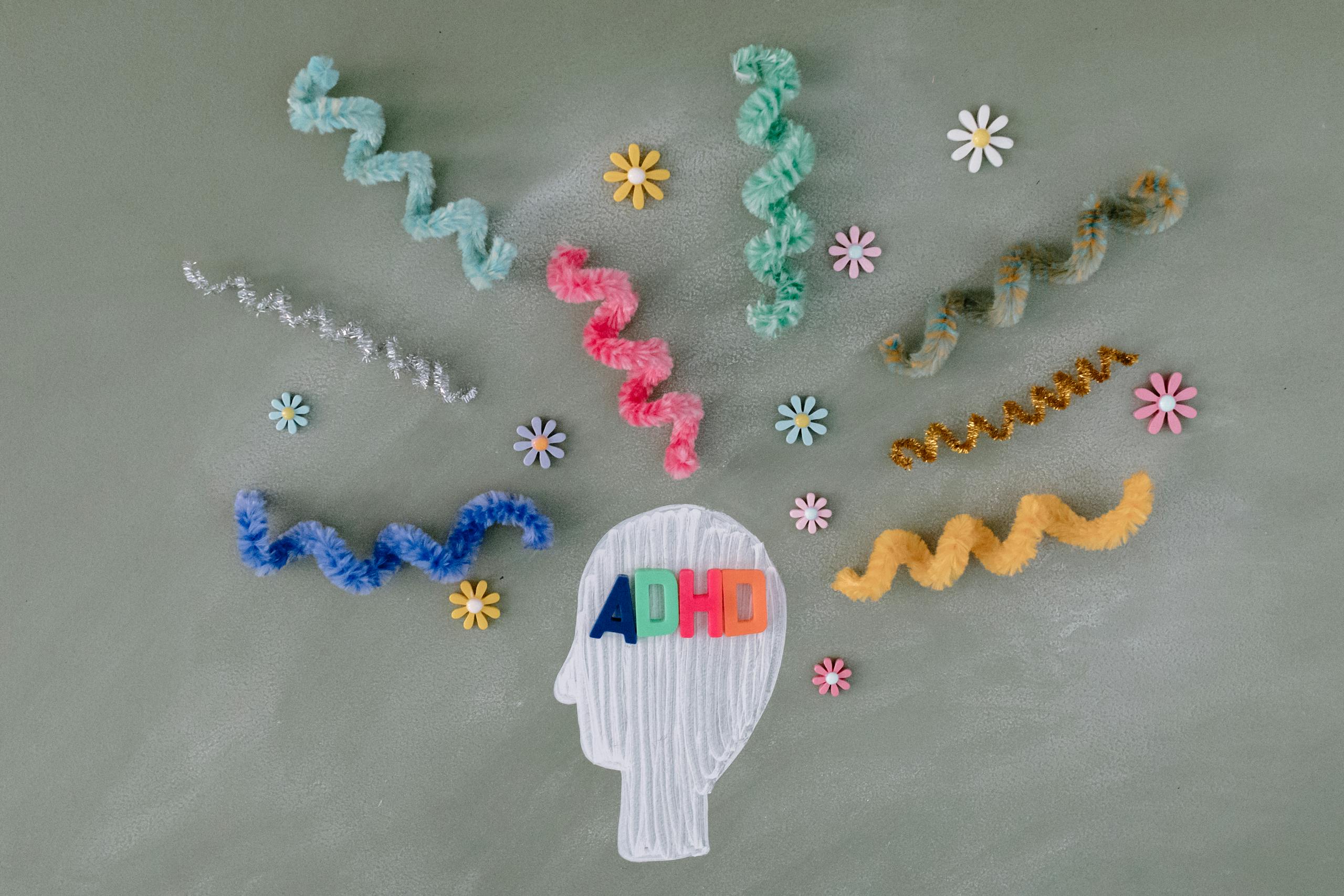 Colorful decorative elements like spiral pipe cleaners and flowers surrounding a drawn silhouette of a head with the letters 'ADHD.'